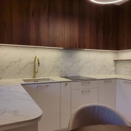 Blenda Studio meble na wymiar - Nowoczesna kuchnia z białymi szafkami, marmurowym blatem i złotą armaturą. Ciemne, drewniane szafki górne i okrągła lampa sufitowa dodają elegancji.