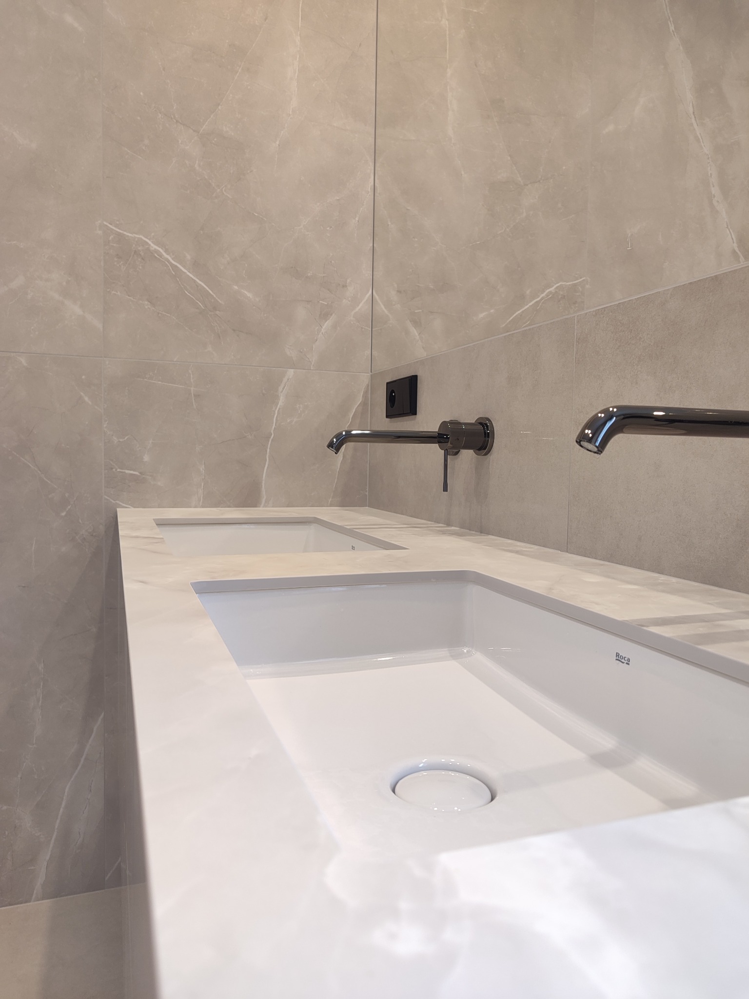 Nowoczesna łazienka z podwójną umywalką nablatową w kolorze białym, z chromowanymi bateriami ściennymi i beżowymi płytkami imitującymi marmur na ścianie.