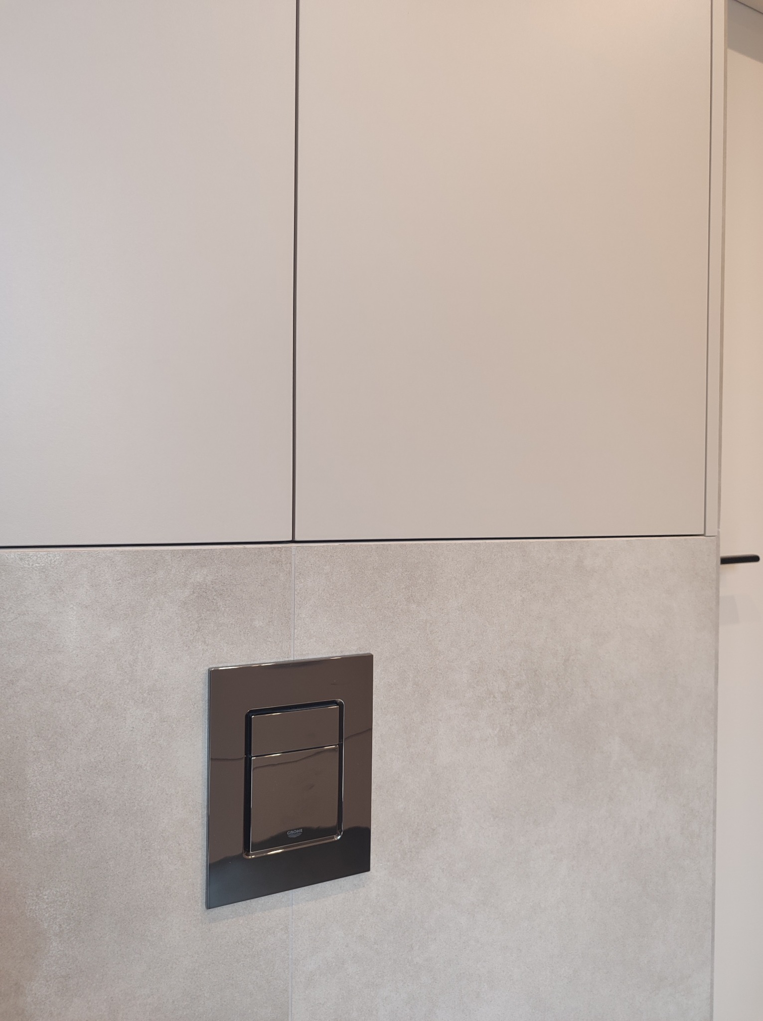 Szare, gładkie fronty szafek kuchennych nad płytkami ściennymi z chromowanym przyciskiem spłuczki WC marki Grohe.