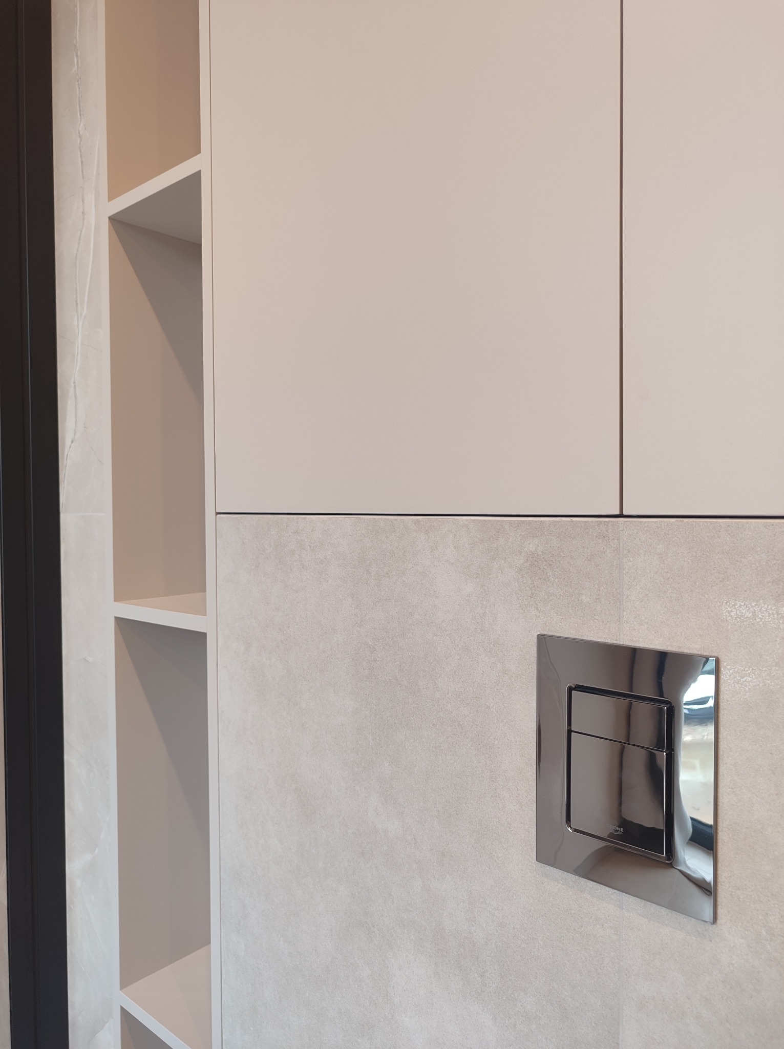 Jasna zabudowa meblowa z otwartymi półkami i chromowanym przyciskiem spłuczki na tle płytek imitujących kamień.