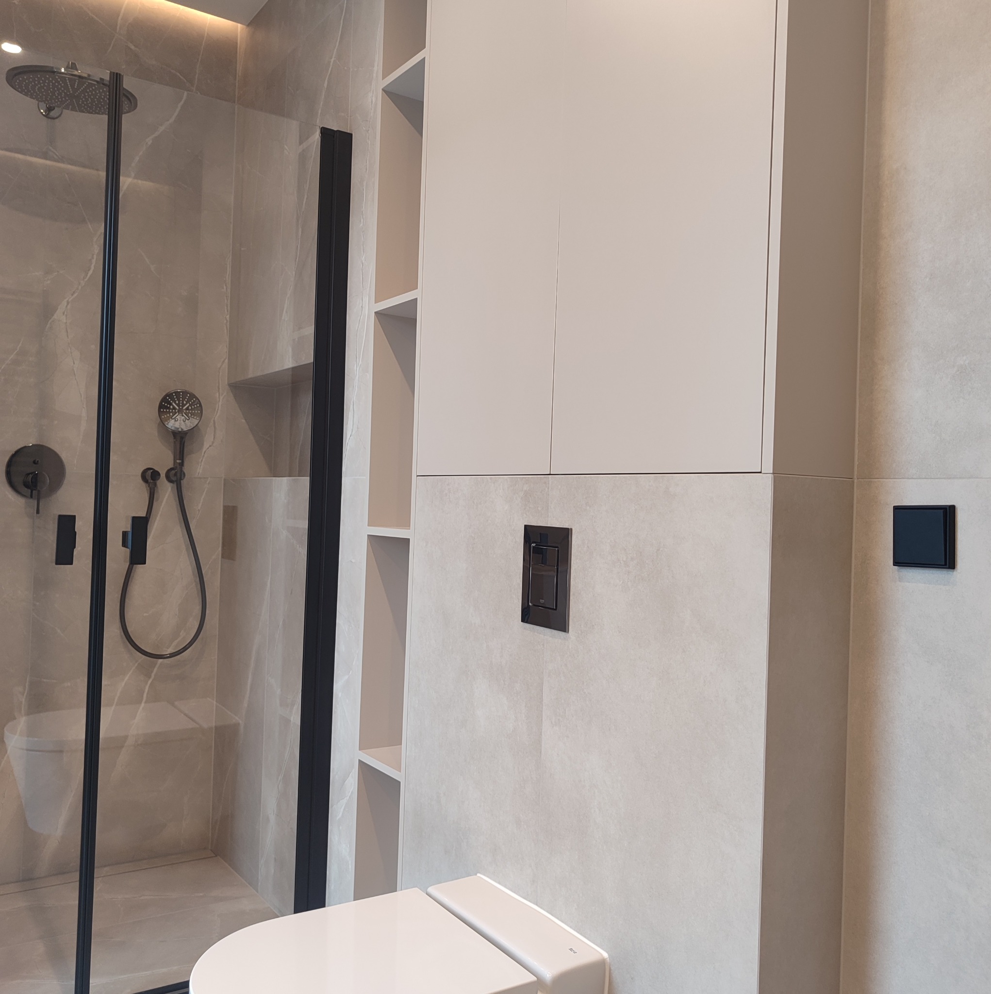 Nowoczesna łazienka z kabiną prysznicową typu walk-in z czarnymi profilami, wiszącą szafką z półkami i toaletą myjącą w beżowej tonacji.