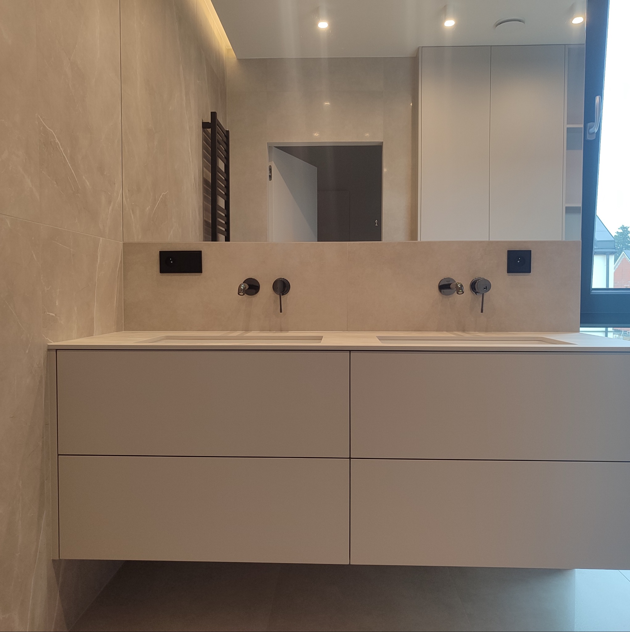 Nowoczesna łazienka w Księżynie z beżową szafką pod umywalkę z czterema szufladami, czarnymi bateriami i gniazdkami, oraz lustrem w zabudowie i grzejnikiem drabinkowym na ścianie wyłożonej płytkami...