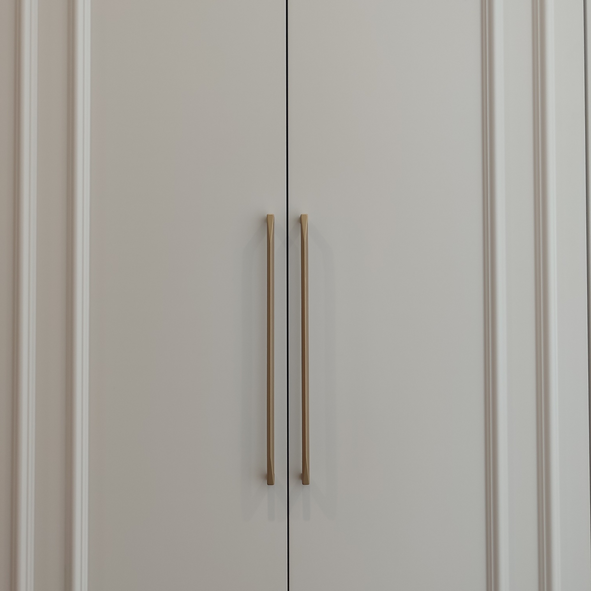 Zamknięte, białe drzwi szafy z frezowanymi krawędziami i długimi, pionowymi, złotymi uchwytami. Minimalistyczny design, symetryczne ujęcie.