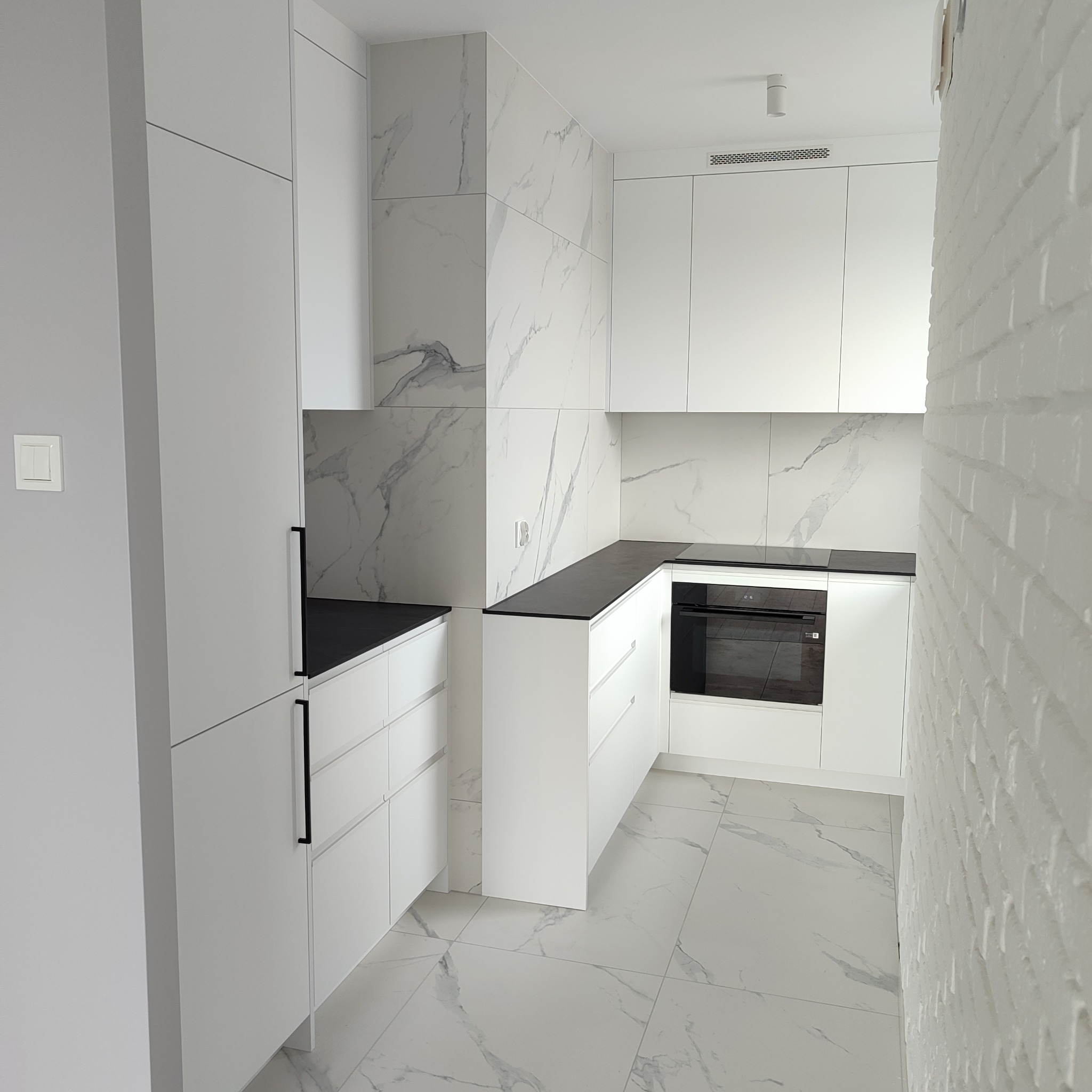 Nowoczesna, biała kuchnia z czarnym blatem i marmurowymi płytkami. Minimalistyczny design, oświetlenie punktowe, widok z perspektywy narożnej.
