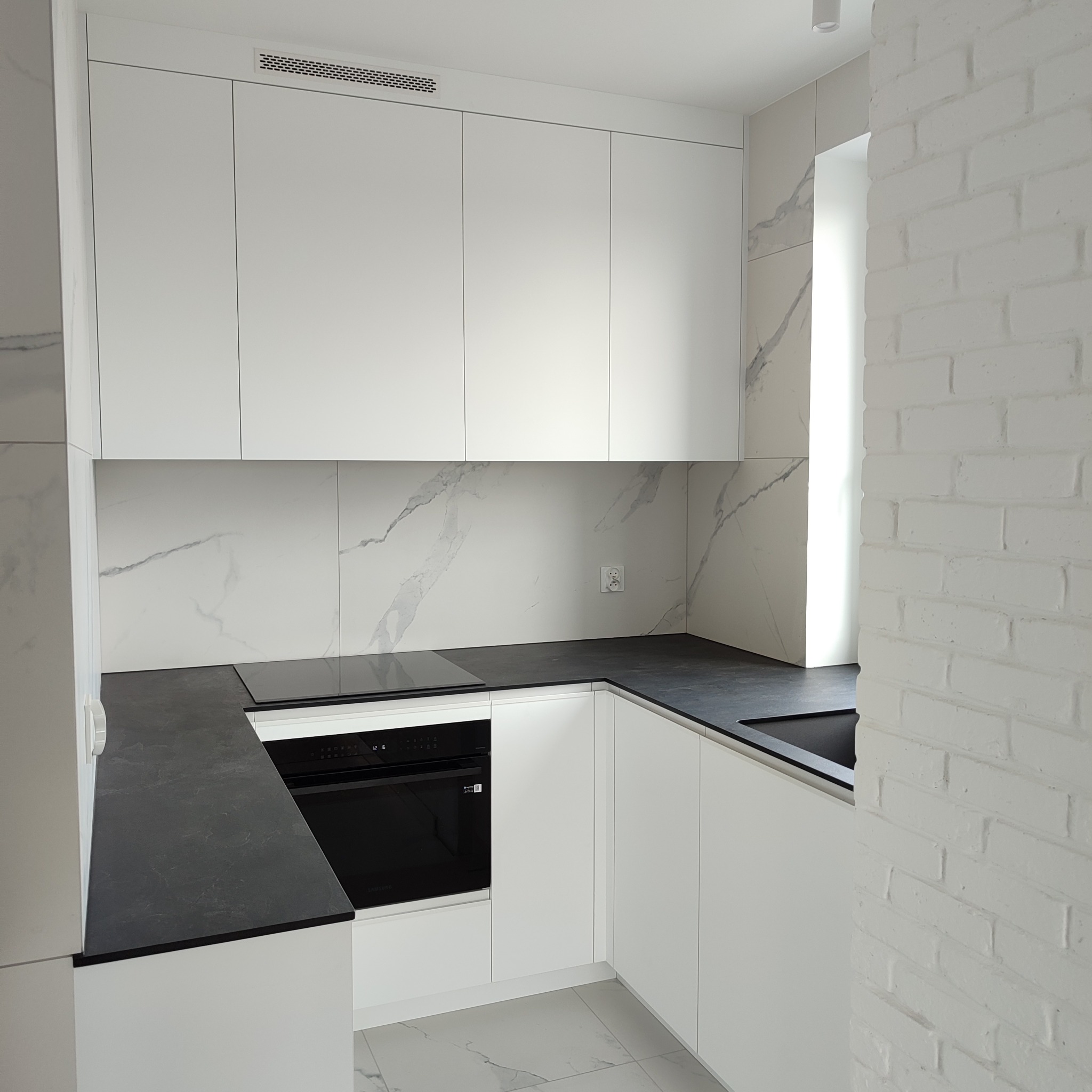 Nowoczesna, biała kuchnia z czarnym blatem i marmurowym backsplashiem. Minimalistyczny design, oświetlenie punktowe, ściana z białej cegły w tle.
