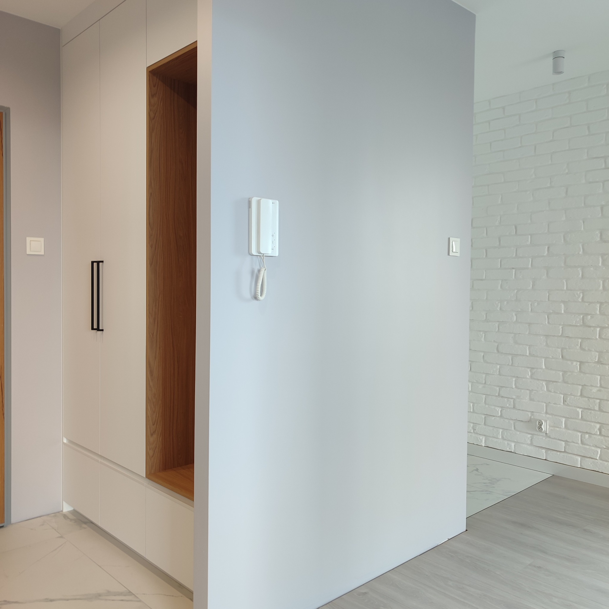 Nowoczesna szafa wnękowa z drewnianymi akcentami i minimalistycznymi uchwytami, wkomponowana w jasne wnętrze z białą cegłą i marmurową podłogą.