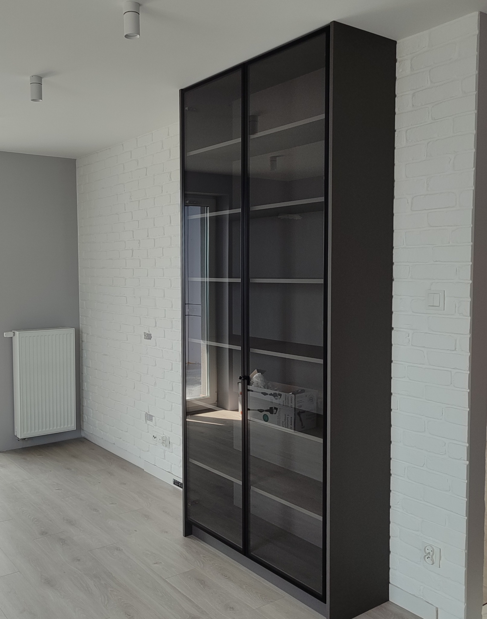 Nowoczesna, wysoka szafa z przesuwnymi, szklanymi drzwiami w ciemnej ramie, na tle białej ściany z cegły i jasnej podłogi. Minimalistyczny design.