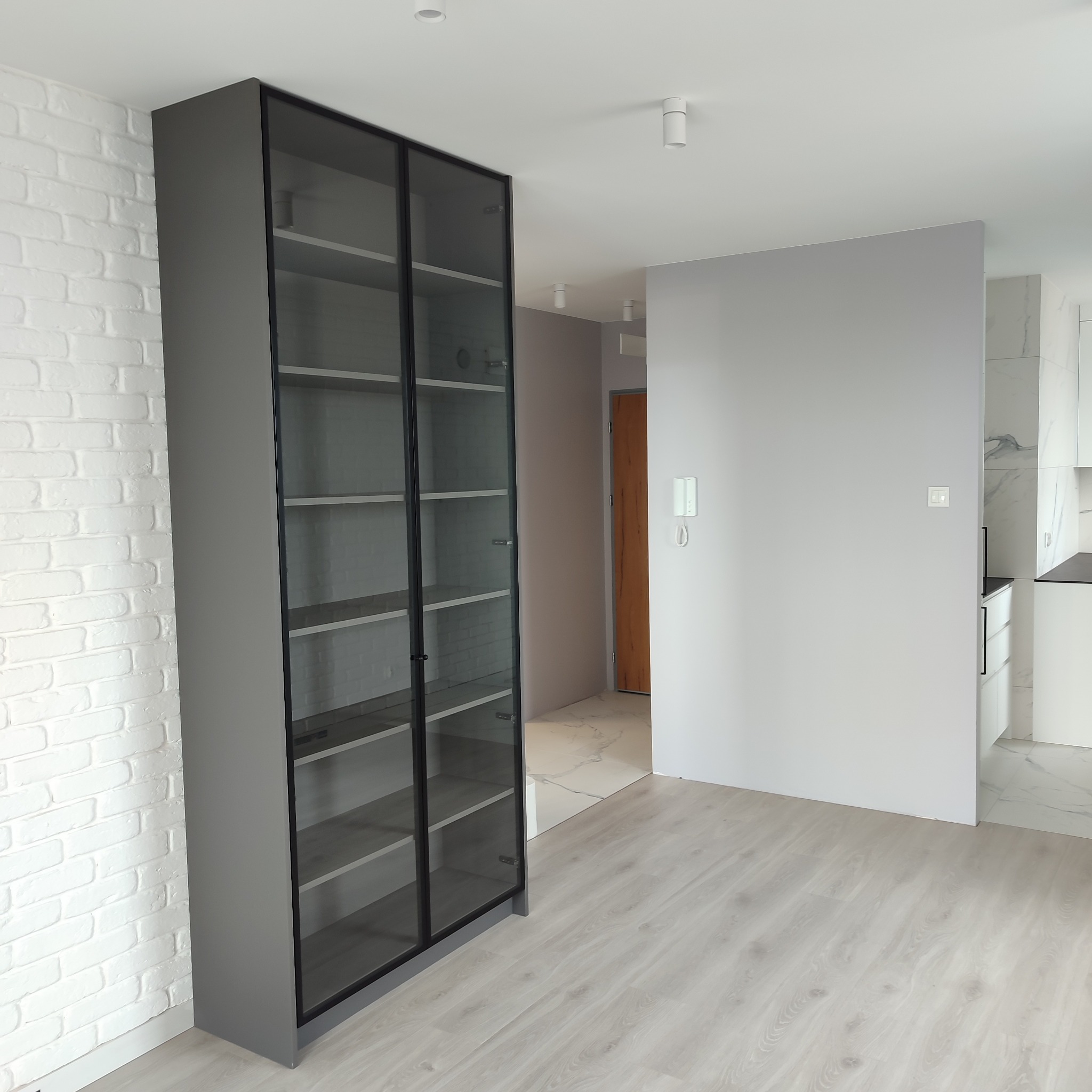 Szara, przeszklona szafa z czarnymi ramami, stojąca na jasnej podłodze, na tle białej ściany z cegły. Widoczne wnętrze mieszkania z elementami kuchni.