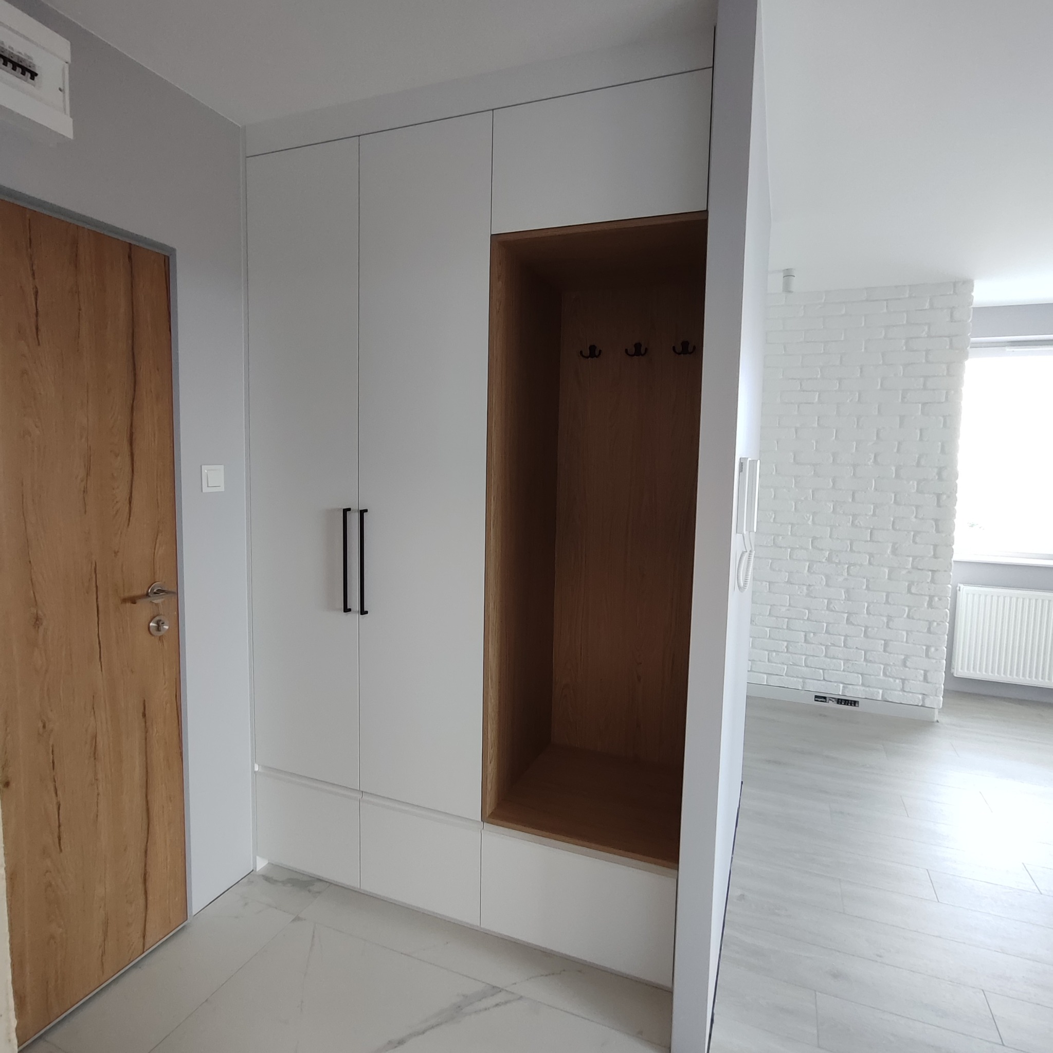 Biała szafa wnękowa z drewnianym akcentem i wieszakami, wkomponowana w przedpokój z drewnianymi drzwiami i białymi cegłami w tle. Minimalistyczny design.