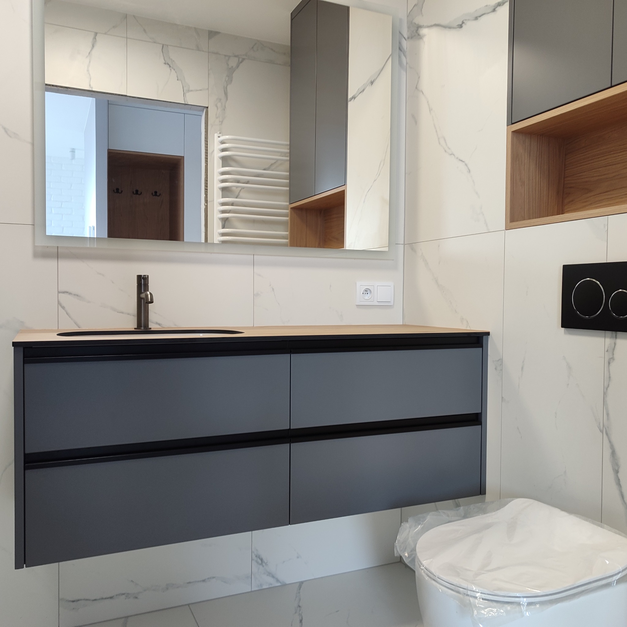 Nowoczesna łazienka z szarą szafką pod umywalką z drewnianym blatem, lustrem z podświetleniem i marmurowymi płytkami. Widoczna toaleta z folią ochronną.