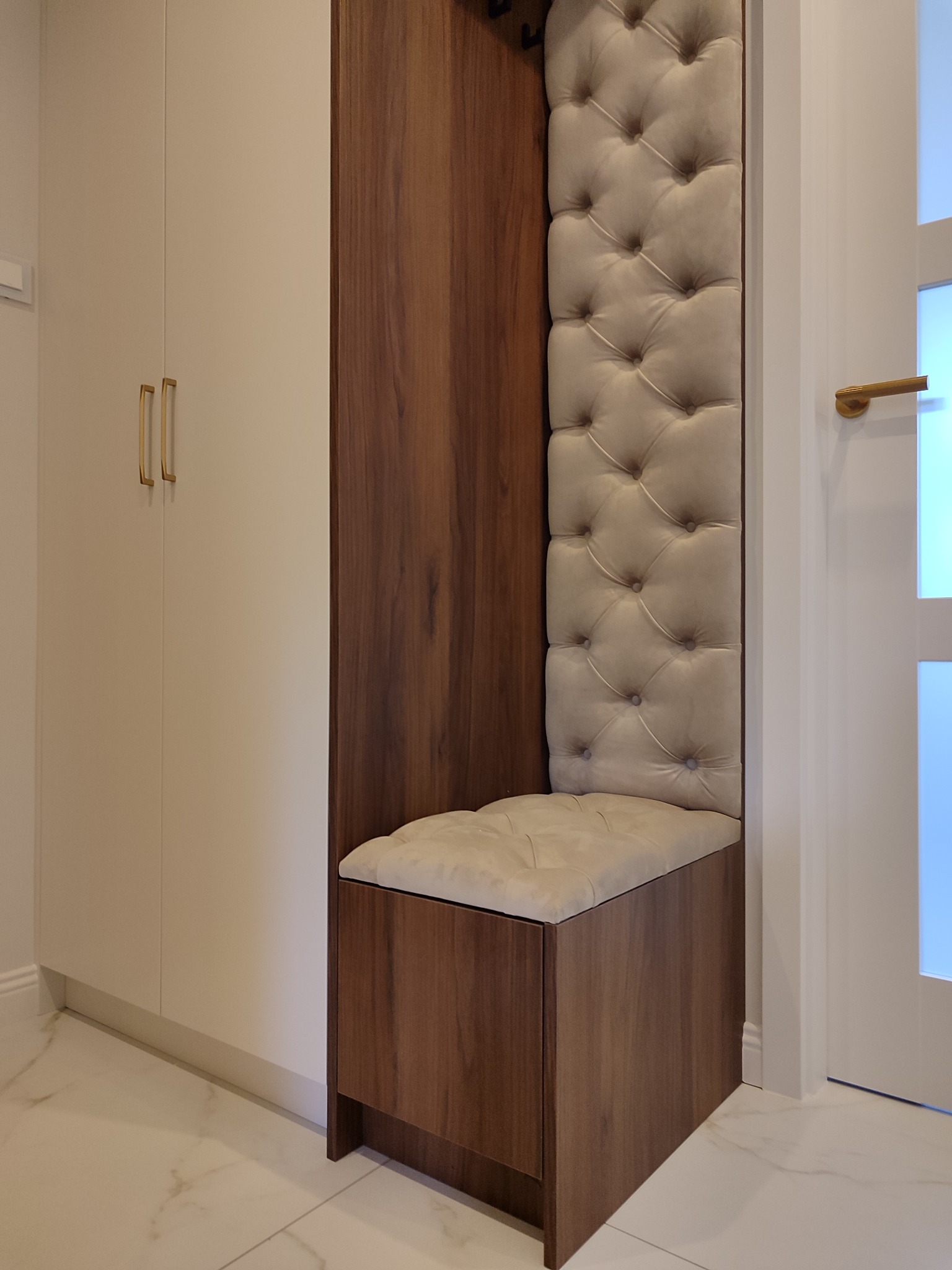 Nowoczesna zabudowa przedpokoju z siedziskiem i tapicerowanym panelem w kolorze beżowym, obok szafa z jasnymi frontami i złotymi uchwytami, marmurowa podłoga.