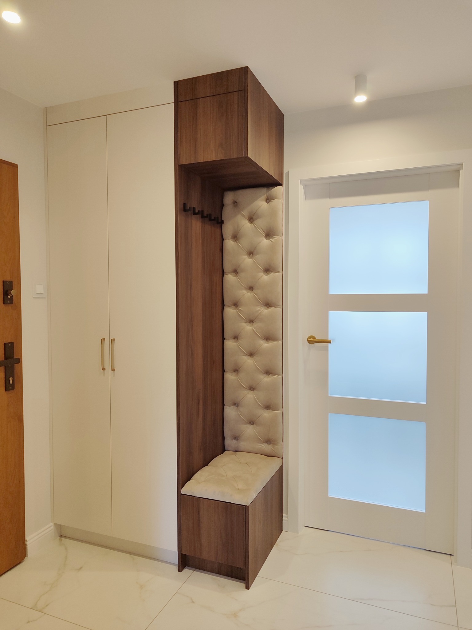 Nowoczesna zabudowa przedpokoju: biała szafa z drewnianym siedziskiem z tapicerowanym oparciem i wieszakami, na tle białych drzwi z przeszkleniami.