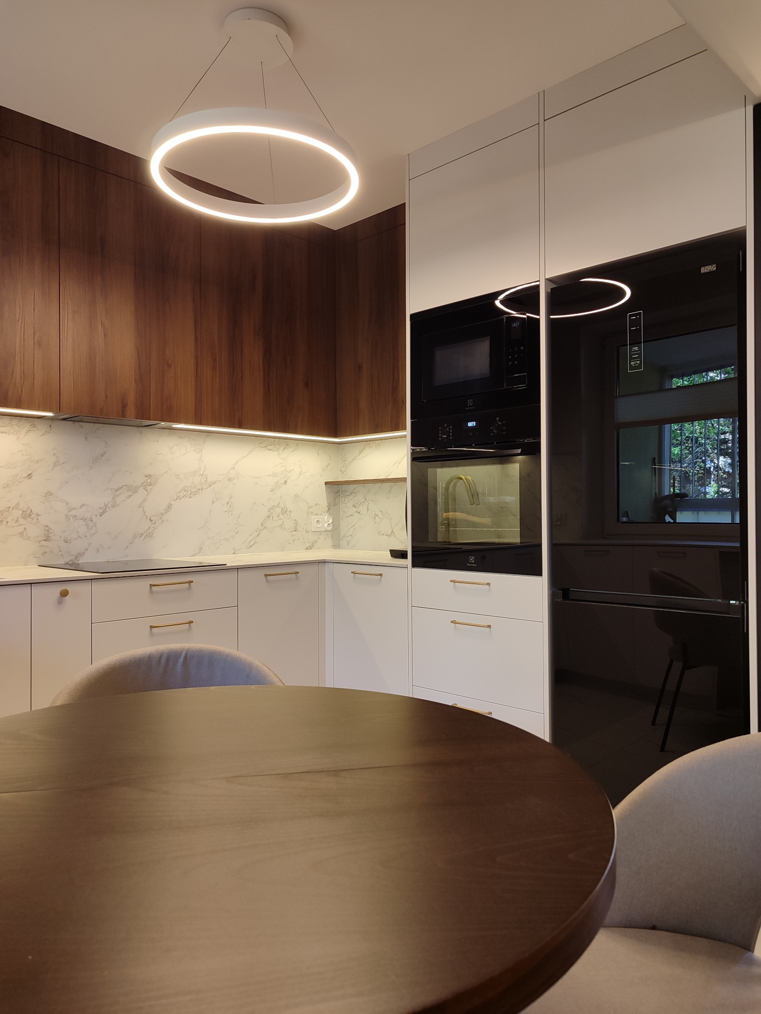 Nowoczesna kuchnia z drewnianymi i białymi szafkami, marmurowym blatem i okrągłym oświetleniem. Widoczny stół z krzesłami. Minimalistyczny design.