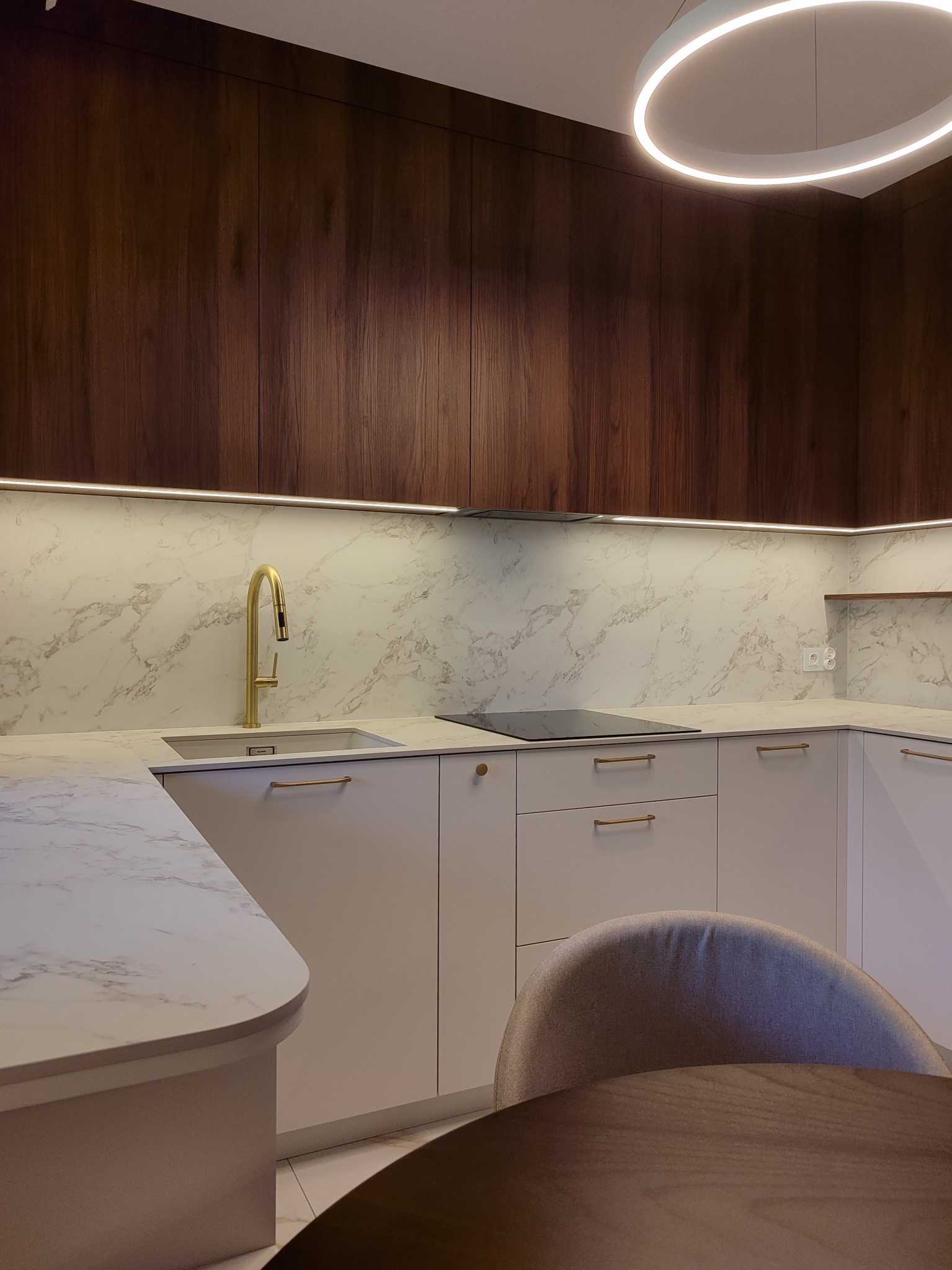 Nowoczesna kuchnia z białymi szafkami, marmurowym blatem i złotą armaturą. Ciemne, drewniane szafki górne i okrągła lampa sufitowa dodają elegancji.
