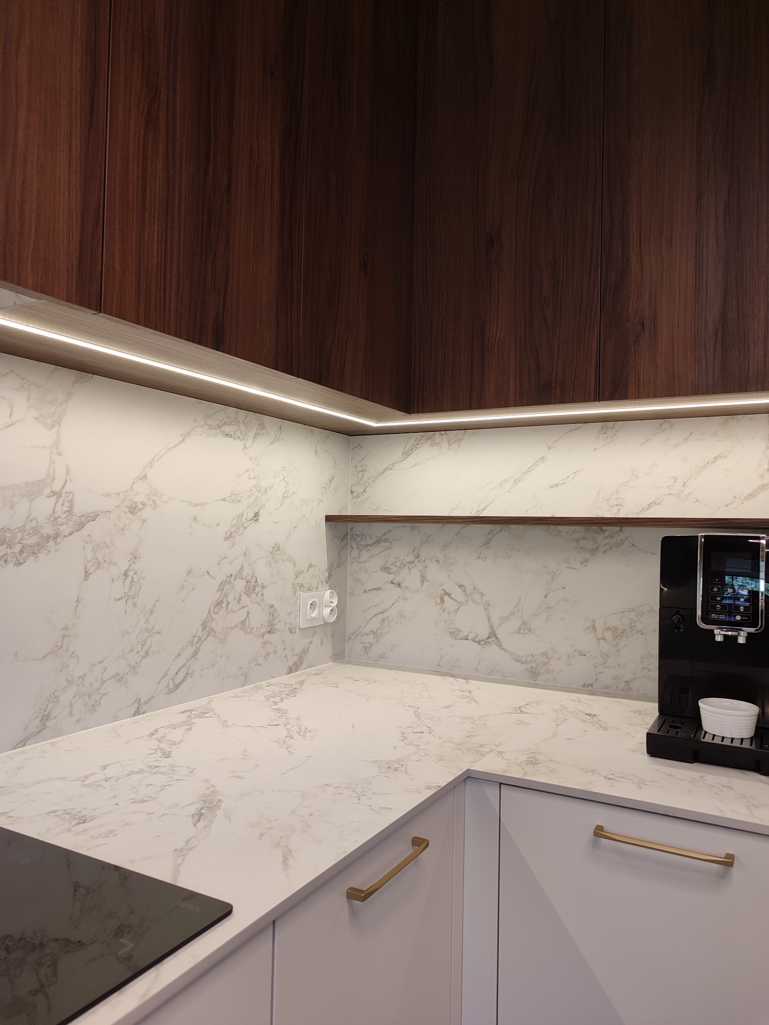 Nowoczesna kuchnia z białymi frontami, złotymi uchwytami i blatem imitującym marmur. Górne szafki z ciemnego drewna, oświetlenie LED pod szafkami.