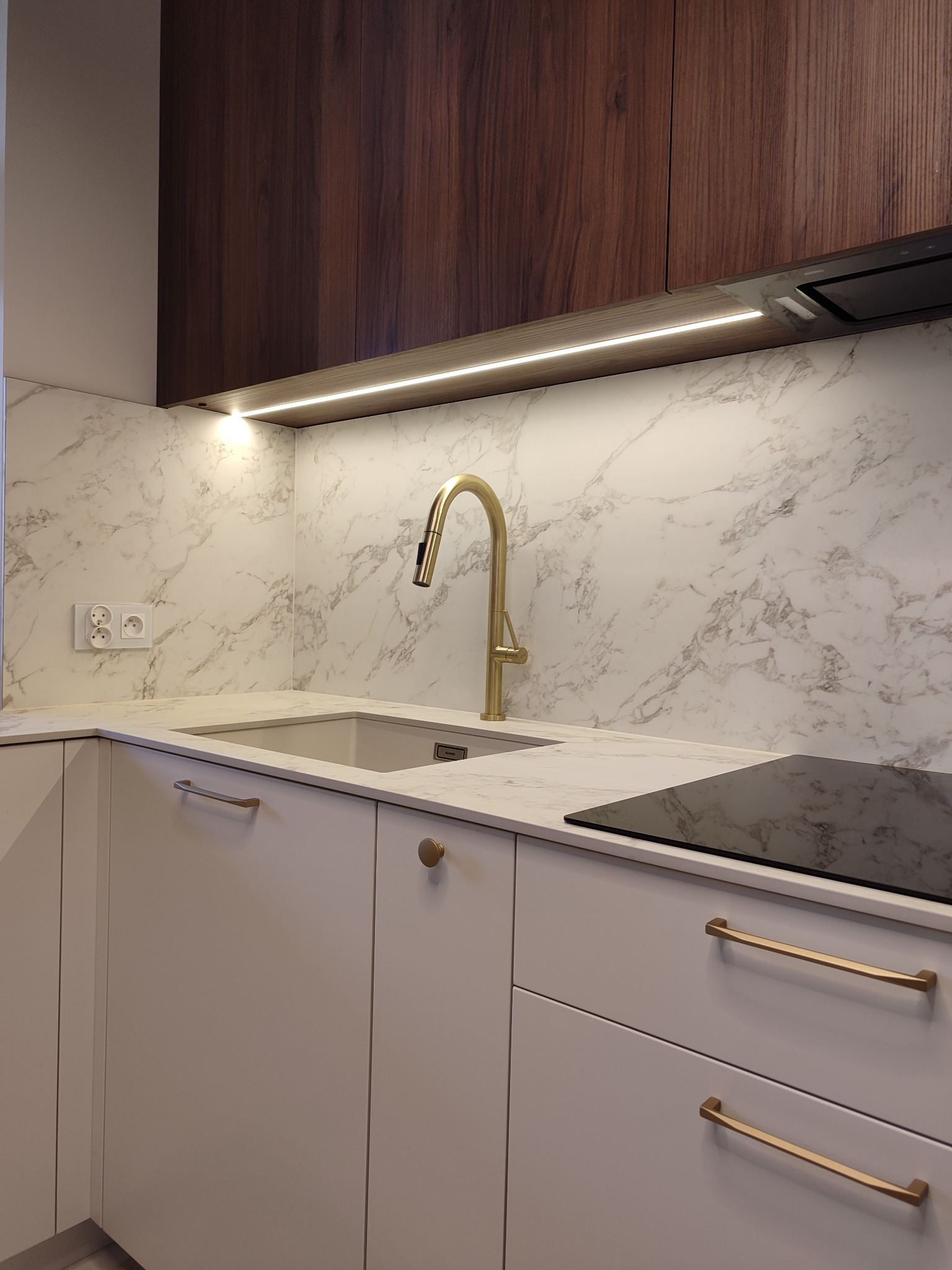 Nowoczesna kuchnia z białymi szafkami, złotą armaturą i blatem imitującym marmur. Górne szafki z ciemnego drewna, podświetlenie LED pod szafkami.