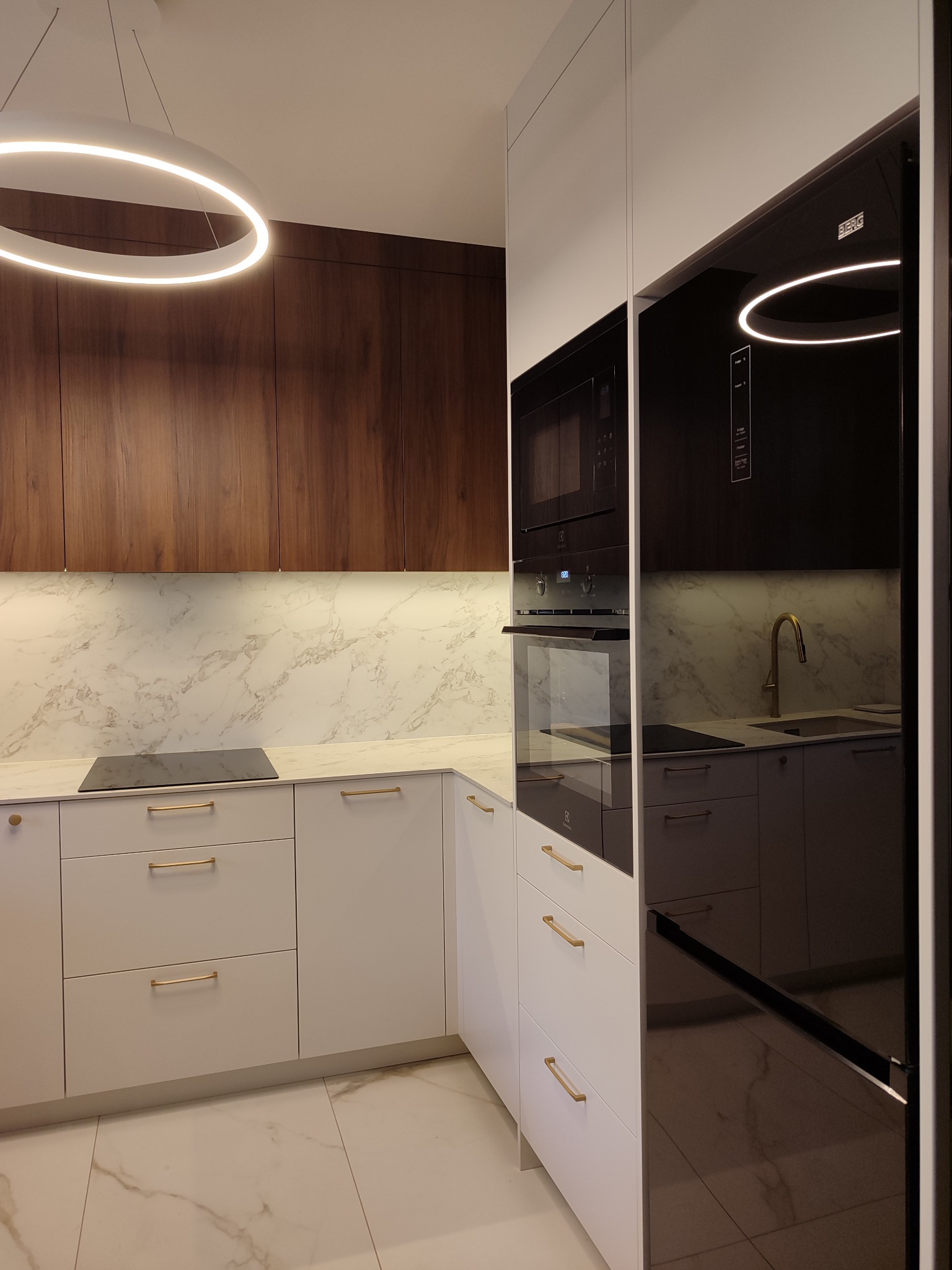 Nowoczesna aranżacja kuchni w Księżynie: białe fronty szafek dolnych z złotymi uchwytami, drewniane szafki górne i marmurowy blat z oświetleniem LED.