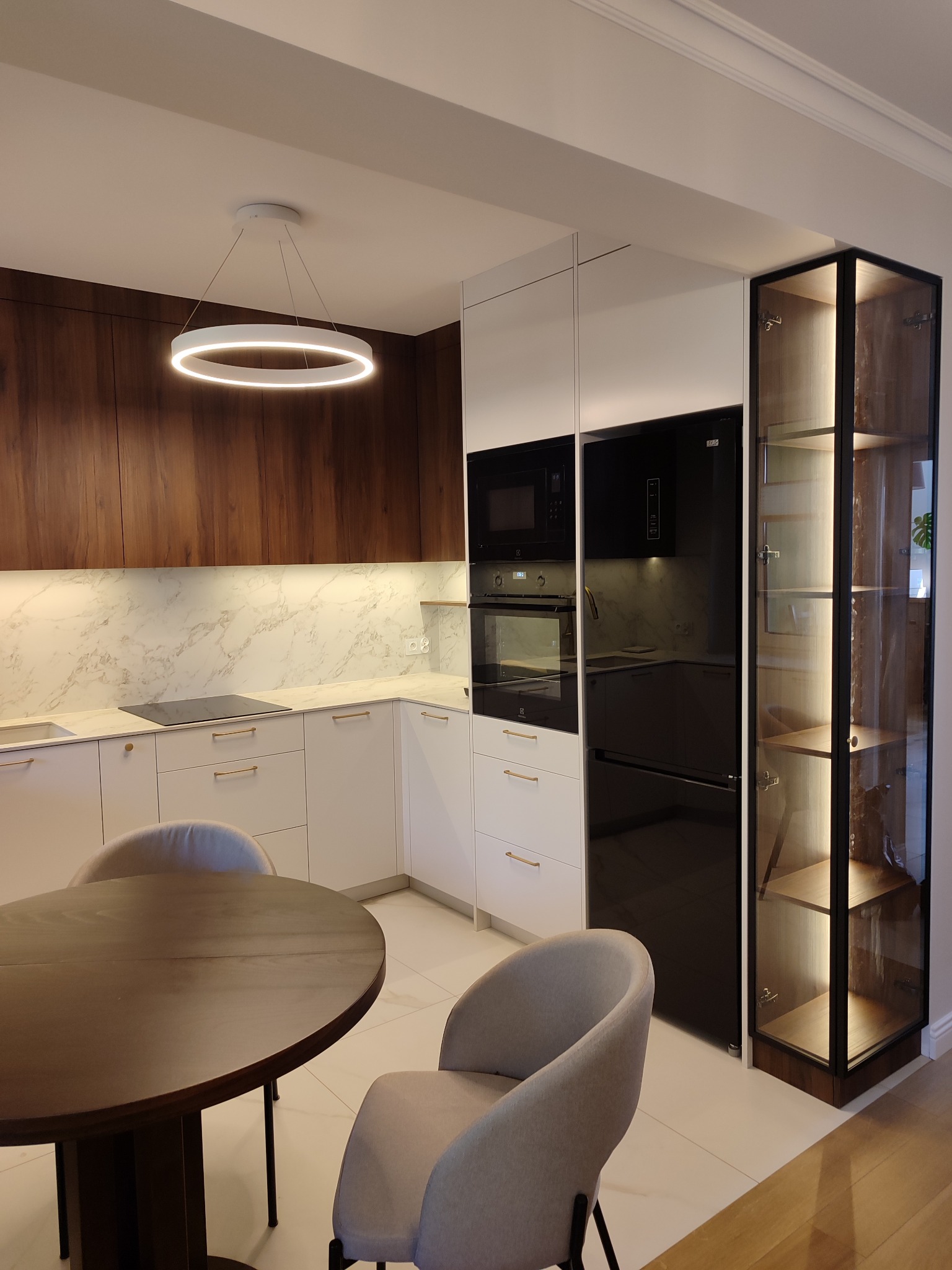 Nowoczesna kuchnia z białymi i drewnianymi frontami, okrągłym stołem i szklaną witryną z podświetleniem LED. Minimalistyczny design i eleganckie wykończenie.