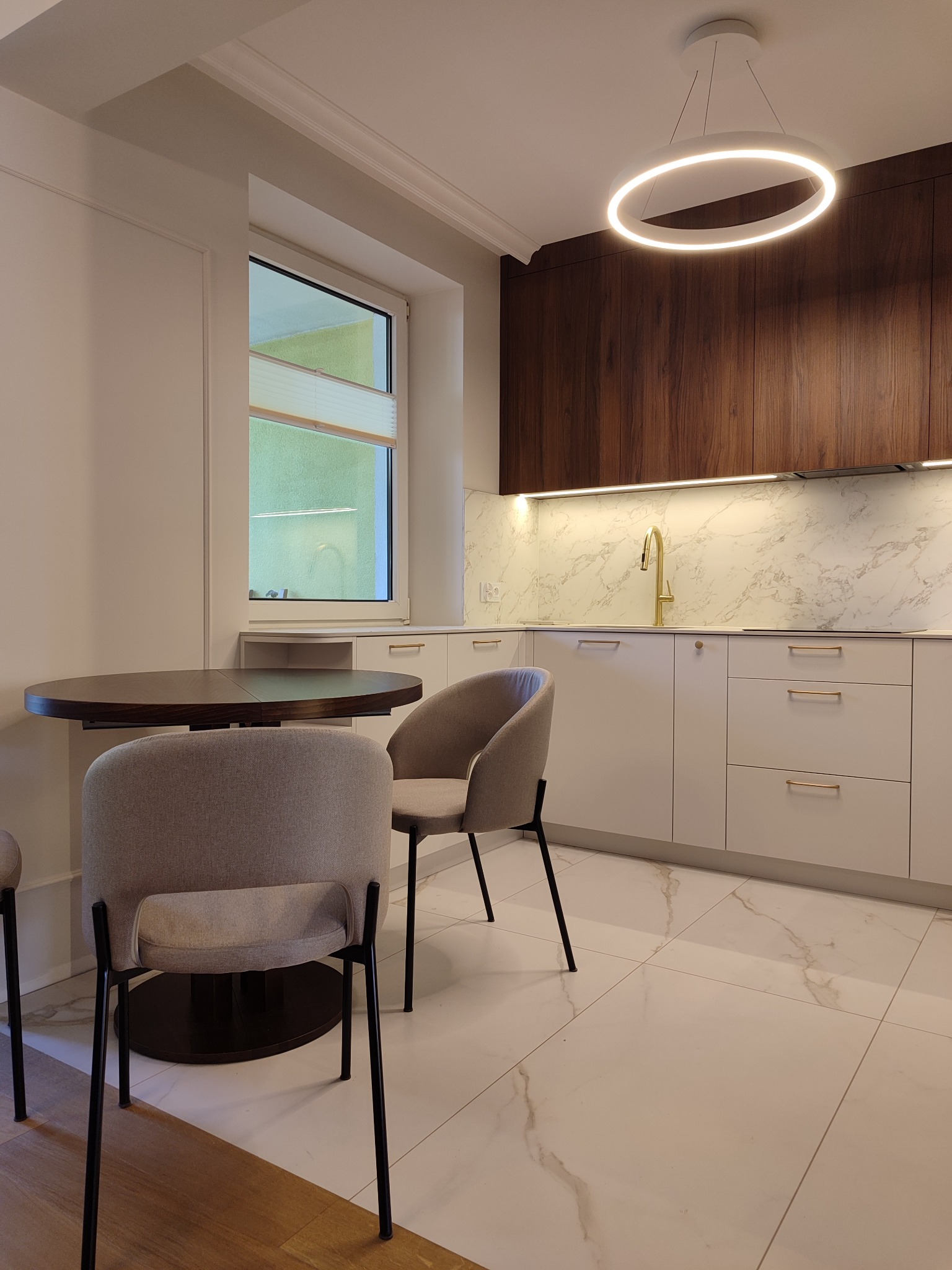 Nowoczesna kuchnia z białymi szafkami, marmurowym blatem i drewnianymi górnymi szafkami. Okrągły stół i krzesła dopełniają aranżację. Oświetlenie LED.