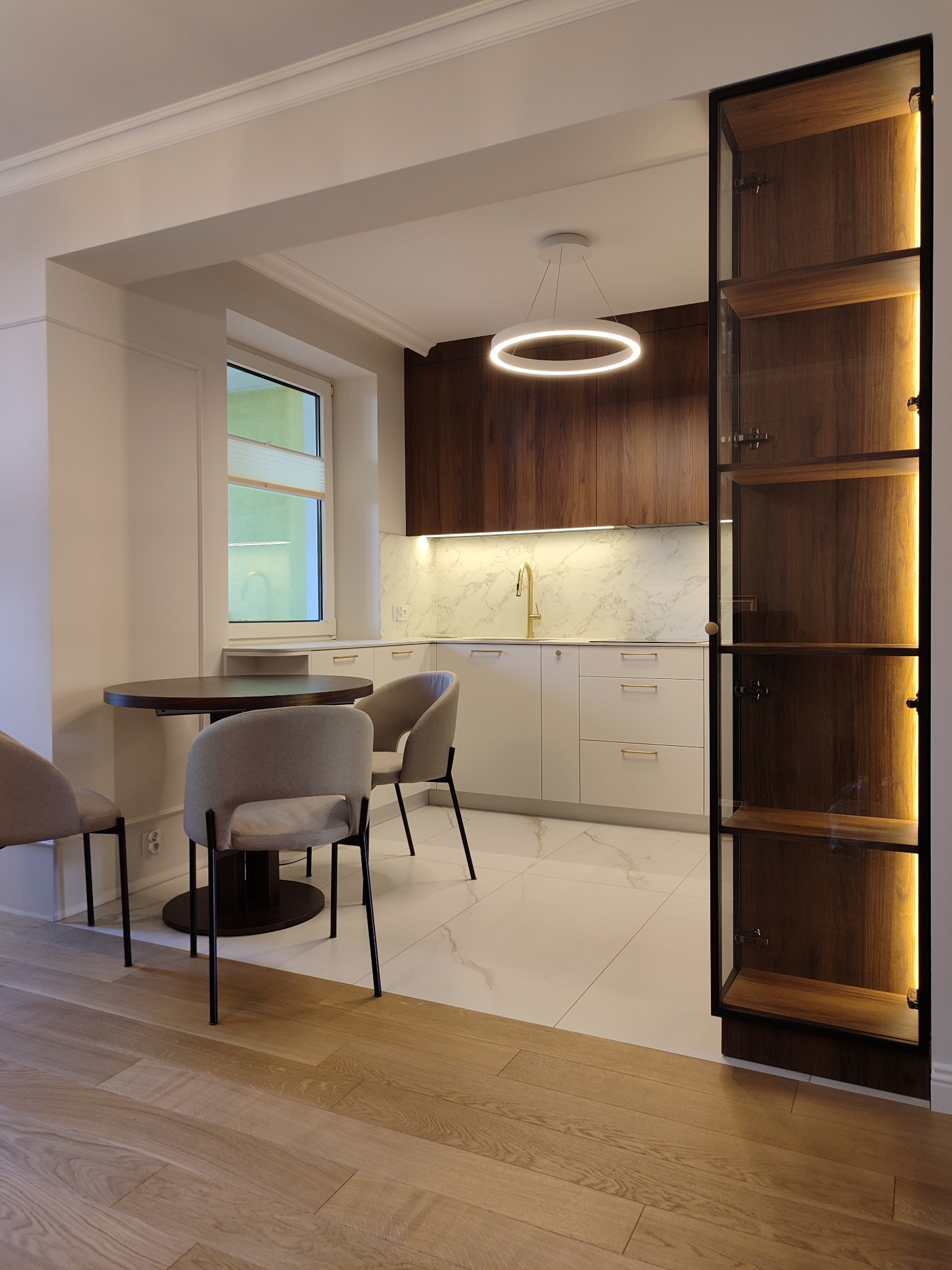 Nowoczesna kuchnia z drewnianymi szafkami, białymi frontami i marmurowym blatem. Okrągły stół z krzesłami i przeszklona witryna z oświetleniem LED.