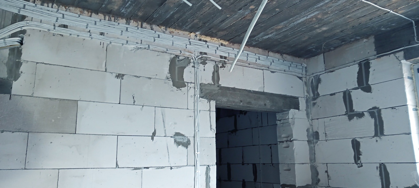 Ściana z betonu komórkowego z zamocowanymi przewodami elektrycznymi w peszlach, ułożonymi równolegle pod sufitem. Widoczne mocowania przewodów i fragment sufitu z desek.