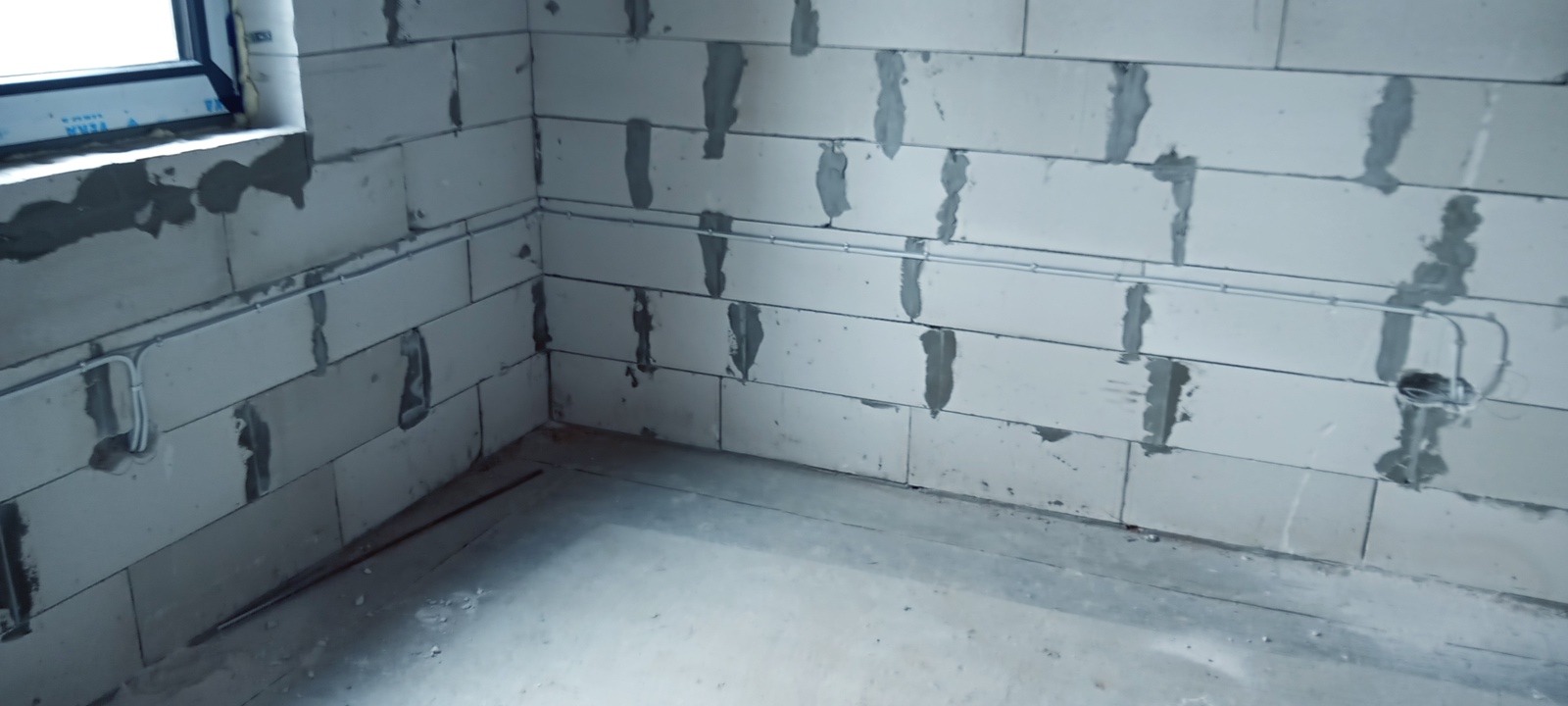 Szare przewody elektryczne poprowadzone po ścianie z bloczków betonowych w surowym wnętrzu, widoczne puszki instalacyjne.