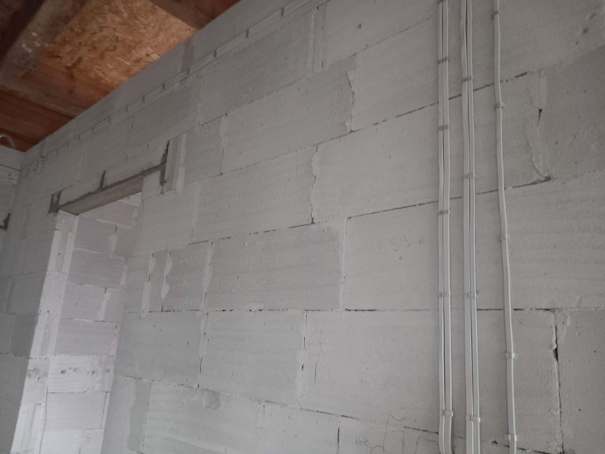 Surowe ściany z betonu komórkowego w trakcie budowy, z widocznymi przewodami elektrycznymi przymocowanymi do ściany, sufit drewniany w tle.