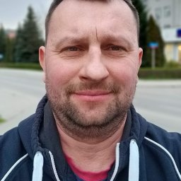 WIKABA Robert Buczek REMONTY I WYKOŃCZENIA - Portret uśmiechniętego mężczyzny w bluzie z kapturem na tle miejskiego krajobrazu z budynkiem i zielenią.