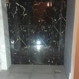 Świeżo ułożone, duże, czarne płytki imitujące marmur z białymi i złotymi żyłami na ścianie łazienki, kontrastują z szarą podłogą z gresu. Widoczne krzyżyki dystansowe między płytkami.