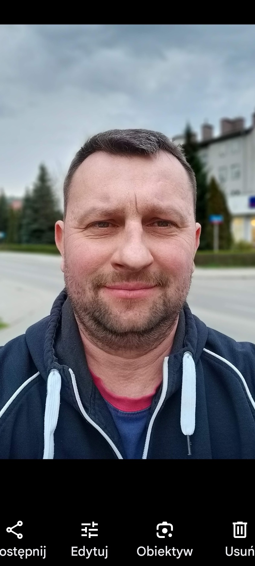 Portret uśmiechniętego mężczyzny w bluzie z kapturem na tle miejskiego krajobrazu z budynkiem i zielenią.