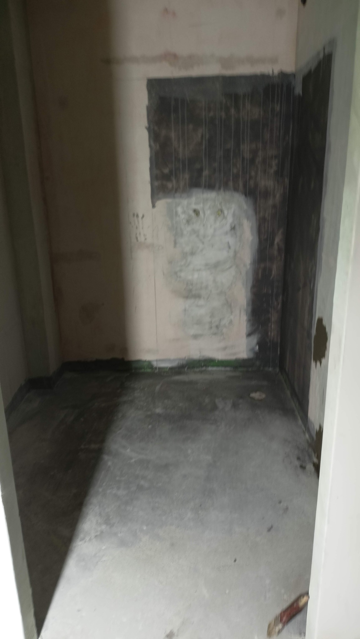 Łazienka w trakcie remontu: ściany częściowo pokryte ciemną masą, widoczne ślady po wcześniejszych pracach, surowy beton na podłodze, brak wykończenia.