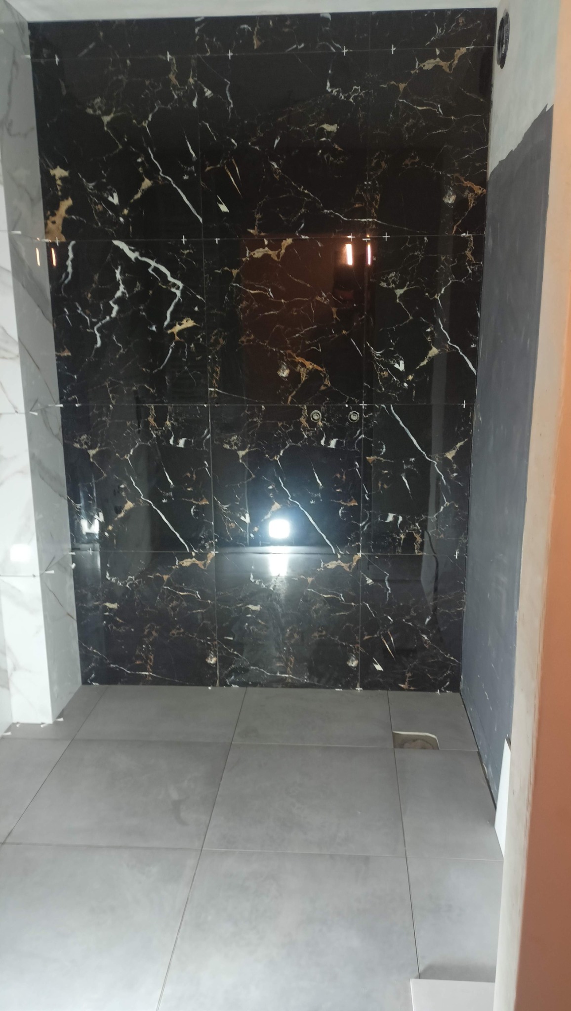 Świeżo ułożone, duże, czarne płytki imitujące marmur z białymi i złotymi żyłami na ścianie łazienki, kontrastują z szarą podłogą z gresu. Widoczne krzyżyki dystansowe między płytkami.