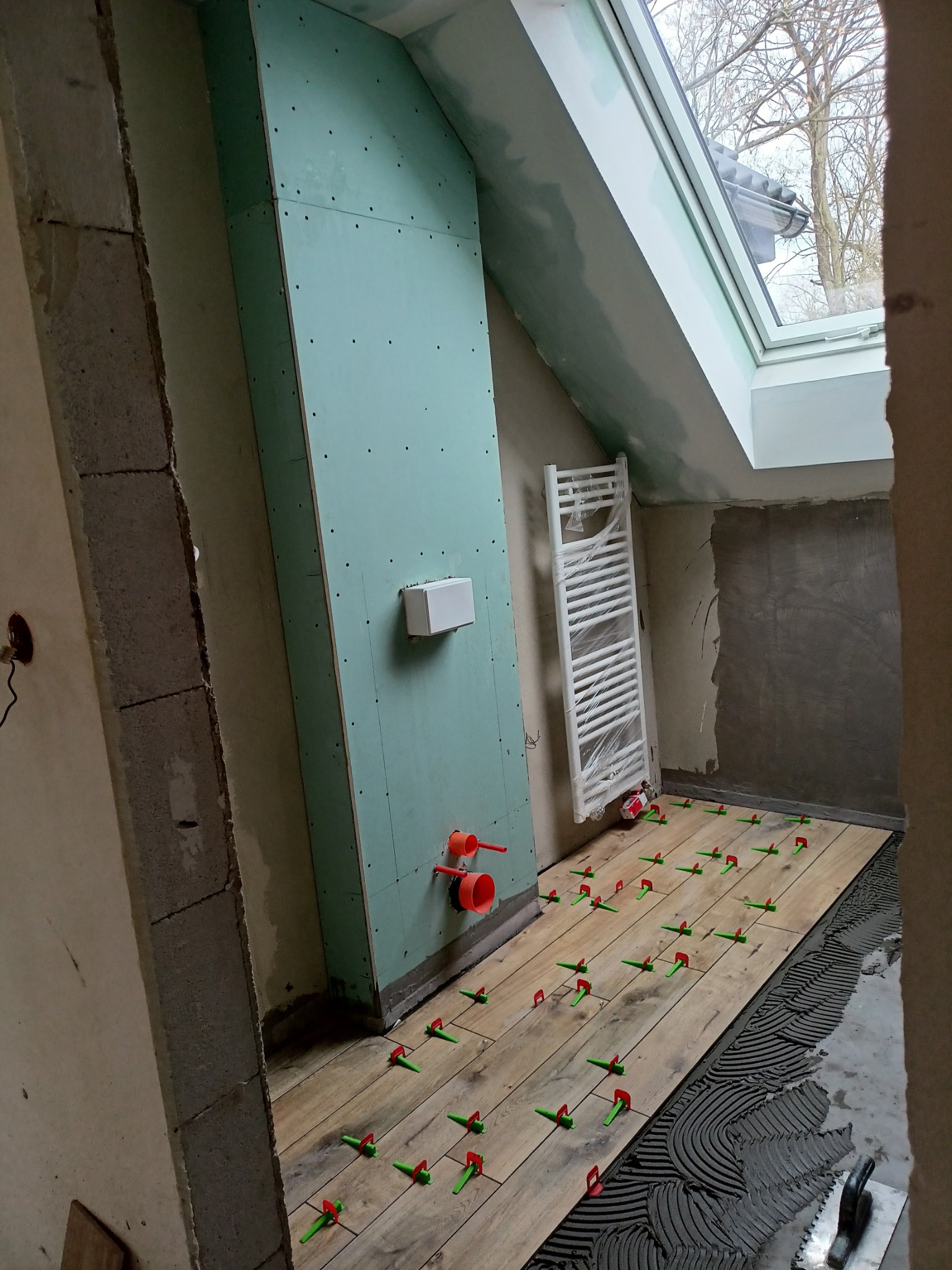 Montaż drewnopodobnych płytek na podłodze w łazience na poddaszu, widoczne plastikowe krzyżyki poziomujące, ściana z płyt gipsowo-kartonowych, grzejnik owinięty folią, okno dachowe.