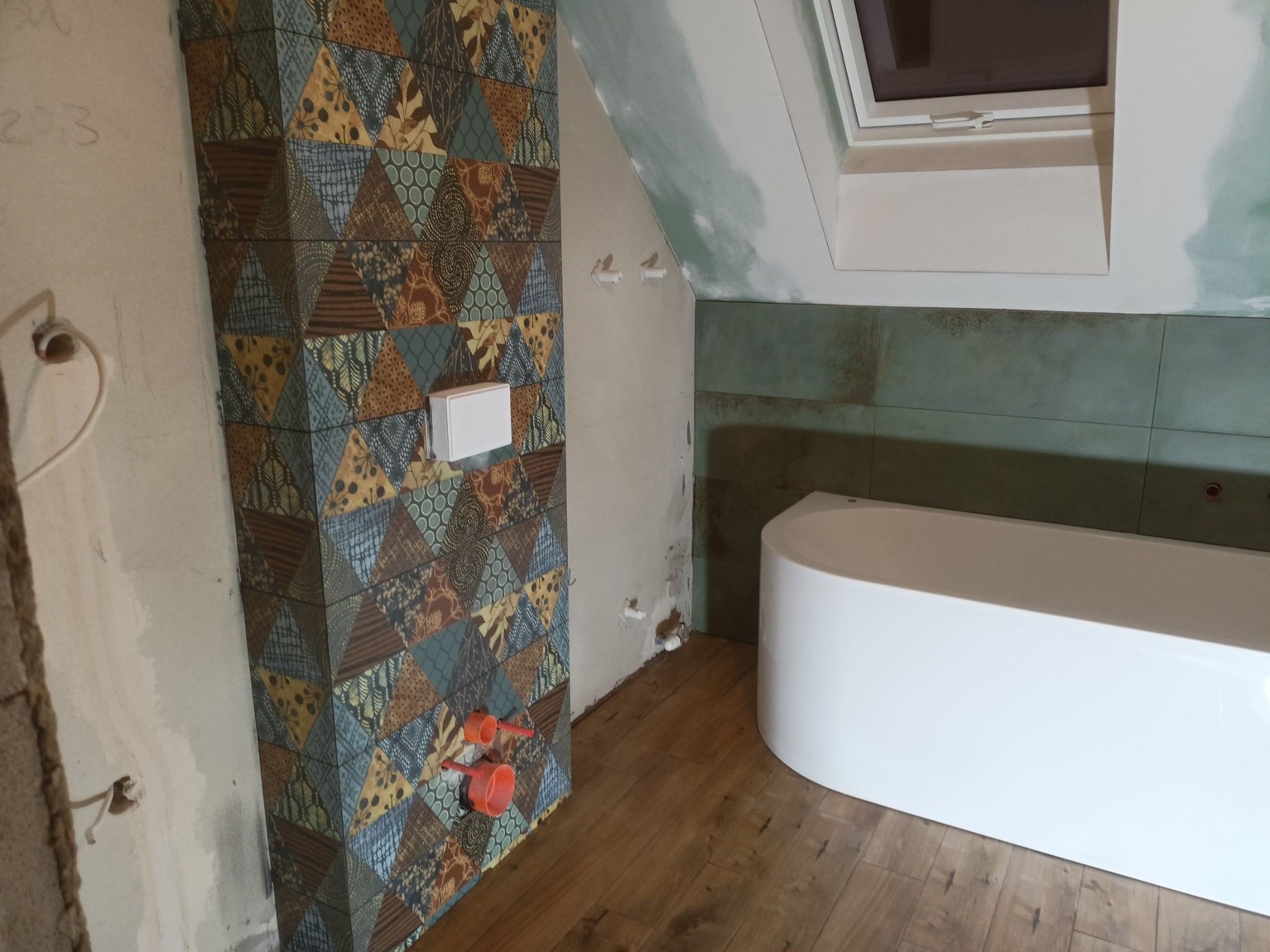 Fragment łazienki w trakcie prac wykończeniowych: ściana z dekoracyjną mozaiką, biała wanna wolnostojąca, drewnopodobna podłoga, widoczne instalacje wodne i okno dachowe.