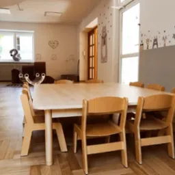 Jasne, przestronne wnętrze sali zabaw z dużym, jasnym drewnianym stołem otoczonym małymi krzesłami, ozdobione ścianami z motywami zwierząt i serduszek.