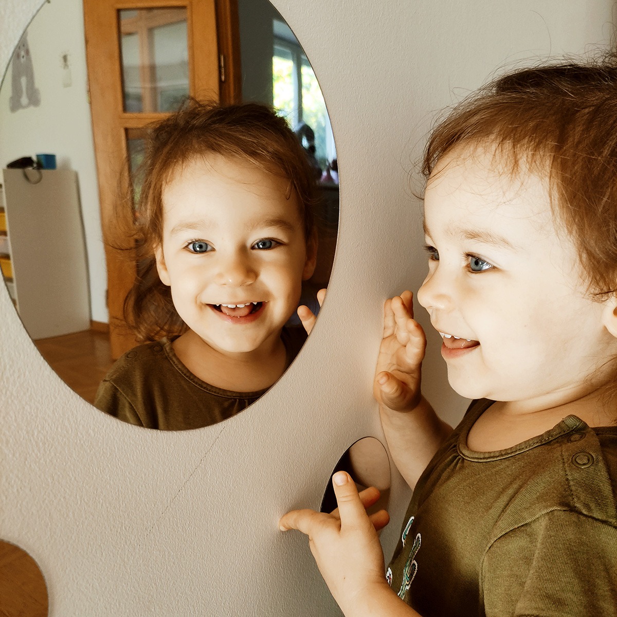 Uśmiechnięta dziewczynka w oliwkowej koszulce, z rozpuszczonymi włosami, bawi się przed lustrem w kształcie księżyca, dotykając jego krawędzi. W tle widoczny pokój dziecięcy z drewnianymi drzwiami.