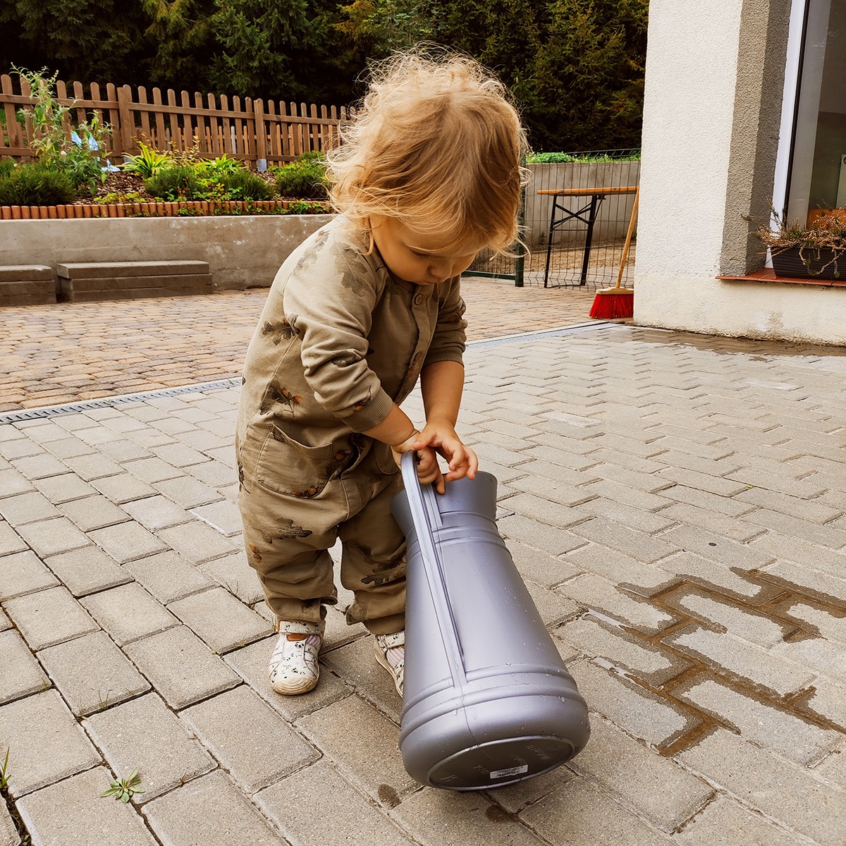 Małe dziecko w kombinezonie z kieszeniami, trzymające dużą, szarą konewkę na brukowanym podwórku, z widocznymi plamami wody.
