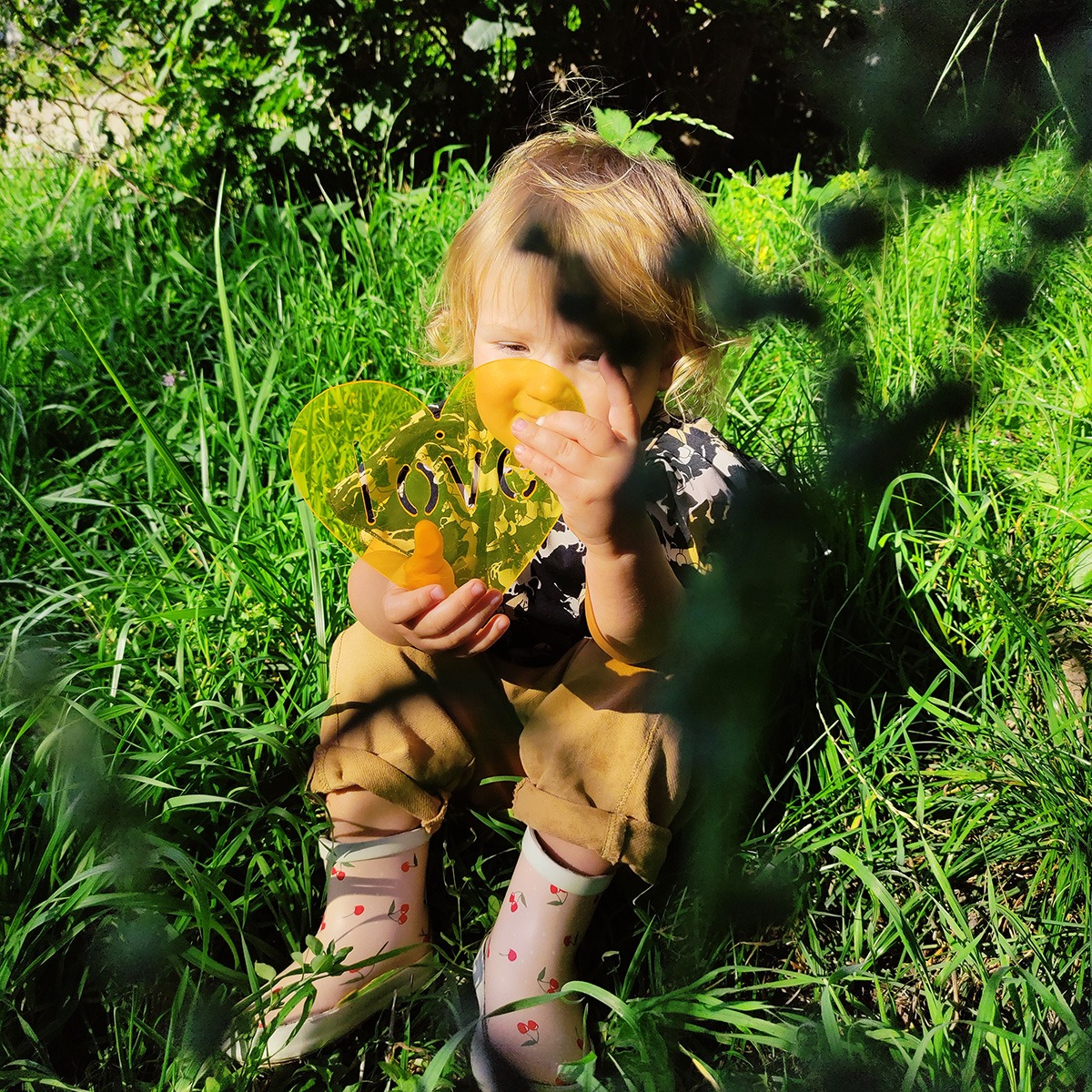 Dziecko siedzi w trawie, trzymając żółte serce z napisem LOVE, ma na sobie kalosze w wisienki.
