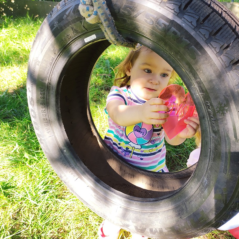 Dziewczynka w pasiastym T-shircie bawi się przez oponę zawieszoną na linie na tle zielonej trawy, trzymając w rękach różowe serduszko.