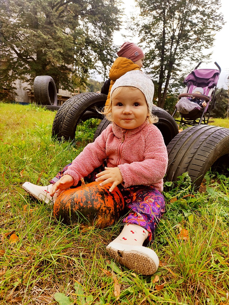 Uśmiechnięta dziewczynka w różowej bluzie i wzorzystych spodniach siedzi na trawie obok pomarańczowej dyni, w tle opony i wózek z dzieckiem.