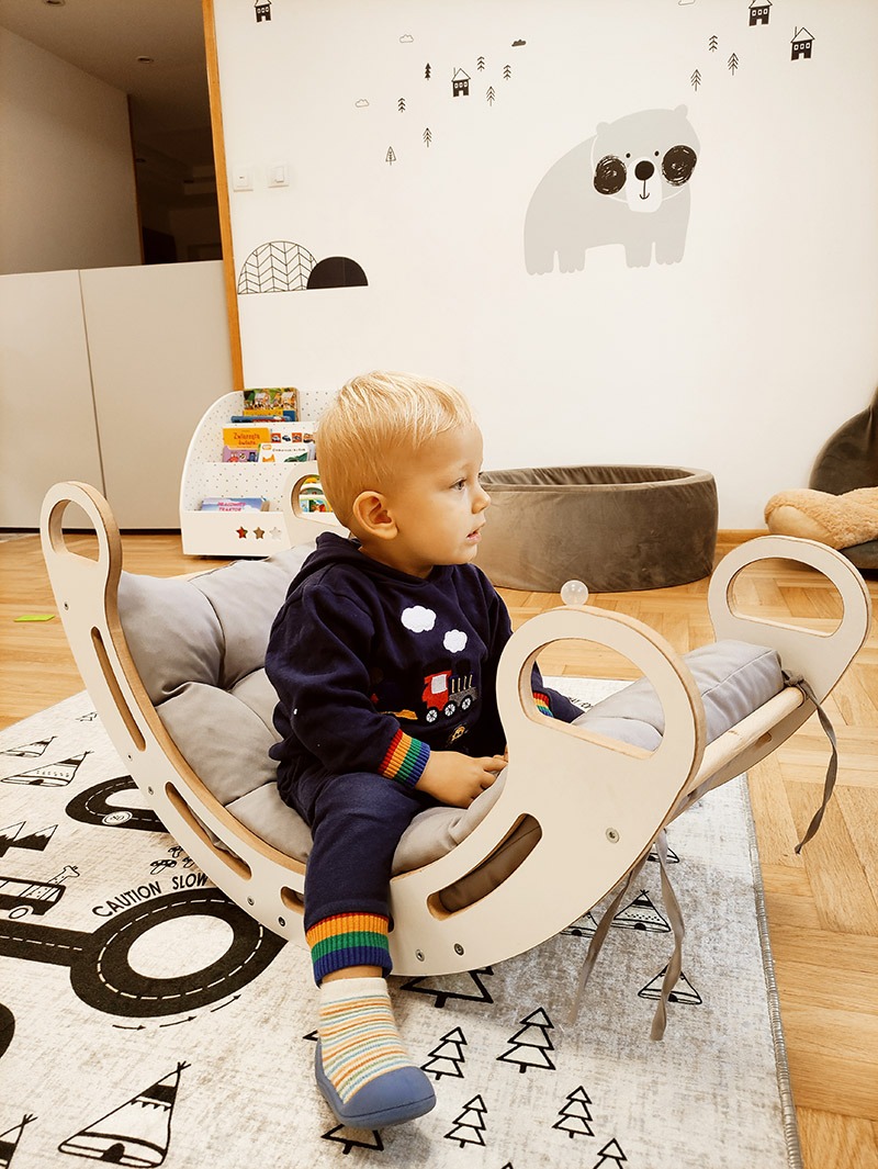 Blondwłosy chłopiec siedzi w drewnianej kołysce z szarymi poduszkami, na dywanie z motywem drogi i wigwamów, w pokoju z dekoracjami ściennymi w stylu skandynawskim.