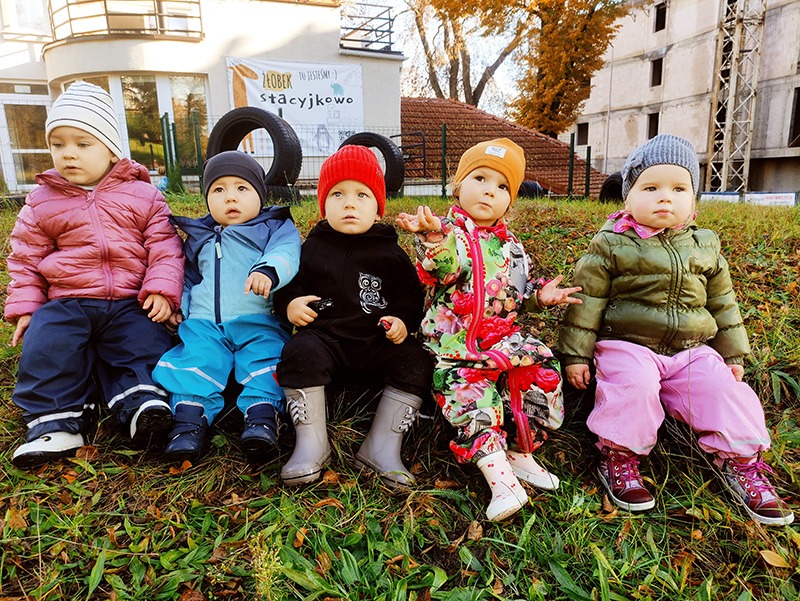 Pięcioro małych dzieci siedzi obok siebie na trawie przed budynkiem żłobka 'Stacyjkowo', z oponami pomalowanymi na czarno w tle.