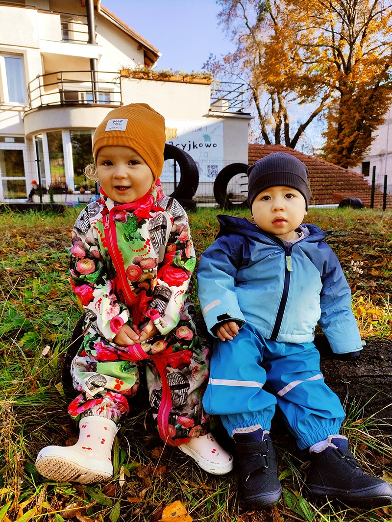 Dwoje małych dzieci w kolorowych kombinezonach siedzi na pniu drzewa na tle budynku z szyldem 'Życzliwe Żłobkowo'.