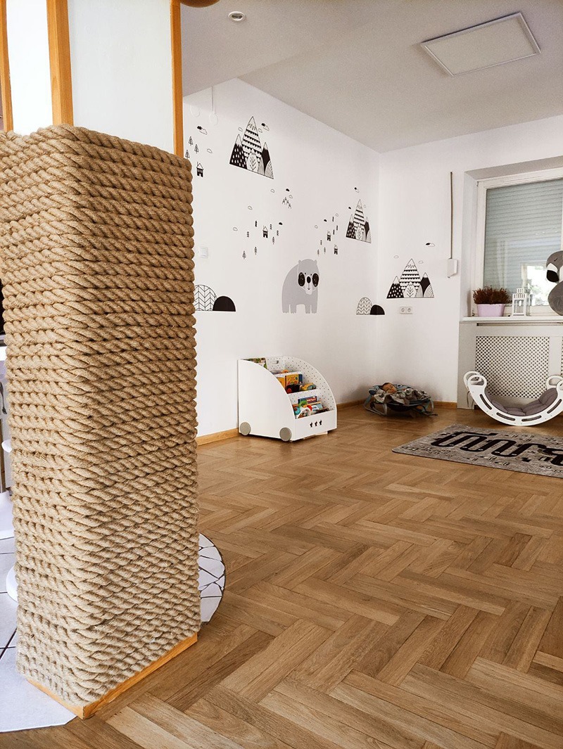 Pokój dziecięcy z drewnianą podłogą w jodełkę, dekoracjami ściennymi z motywem gór i niedźwiedzia, regałem na kółkach z książkami i drapakiem dla kota obitym sznurkiem jutowym.