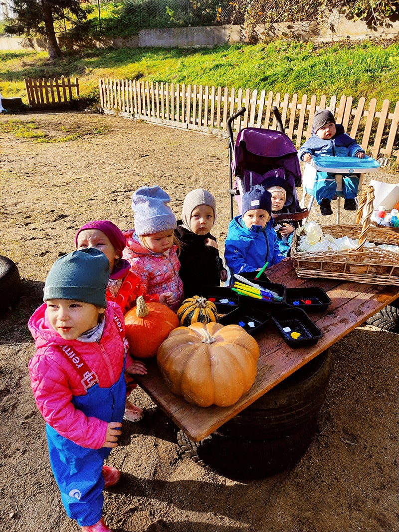 Grupa dzieci w Gdyni, w różnokolorowych ubraniach i czapkach, zgromadzona wokół drewnianego stołu z dyniami i farbami, część siedzi w wózku lub krzesełku do karmienia na zewnątrz.