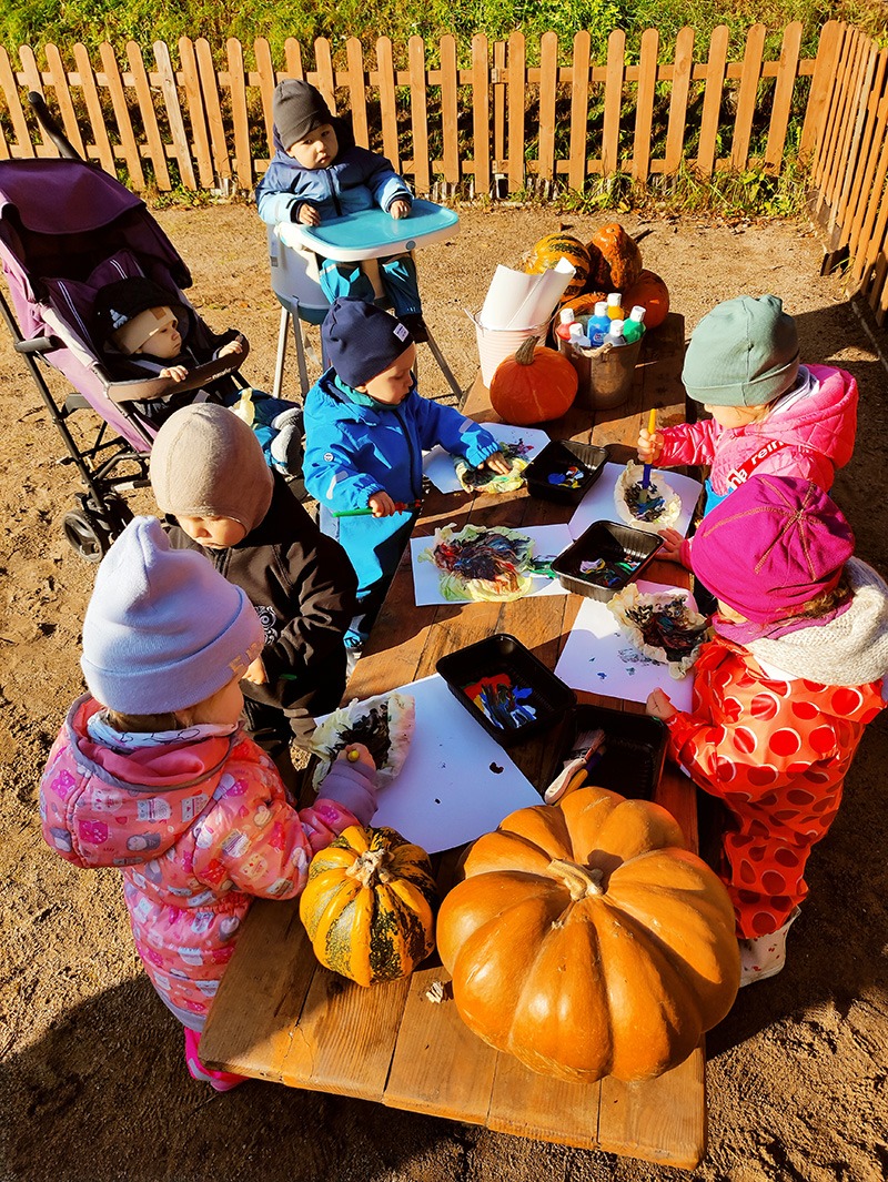 Grupa małych dzieci maluje farbami na papierze przy drewnianym stole na zewnątrz, z dyniami jako dekoracją, jedno dziecko siedzi w krzesełku do karmienia, a drugie w wózku.