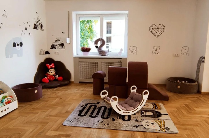 Jasny pokój zabaw dla dzieci z drewnianą podłogą w jodełkę, miękkimi klockami, suchym basenem z kulkami, huśtawką w kształcie łuku, dywanem z motywem toru wyścigowego i naklejkami na ścianach...