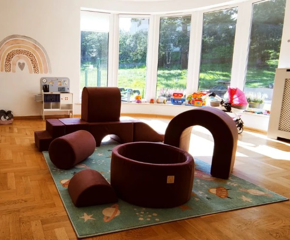 Pokój zabaw dla dzieci z miękkimi, brązowymi klockami piankowymi, basenem z kulkami, kuchnią dla dzieci i tęczową naklejką na ścianie, oświetlony przez duże okno.