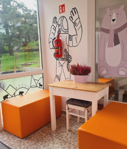 Wnętrze żłobka z pomarańczowymi pufami, białym stolikiem z kwiatami i rysunkiem astronauty z gaśnicą na ścianie oraz dekoracją z motywem niedźwiedzia na szybie.