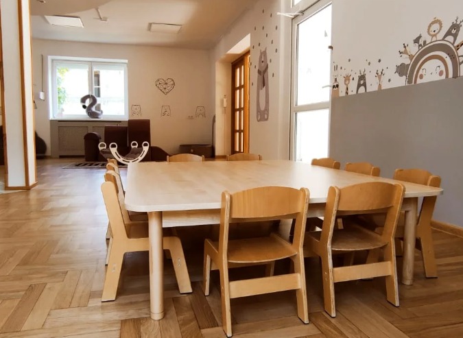 Jasne, przestronne wnętrze sali zabaw z dużym, jasnym drewnianym stołem otoczonym małymi krzesłami, ozdobione ścianami z motywami zwierząt i serduszek.